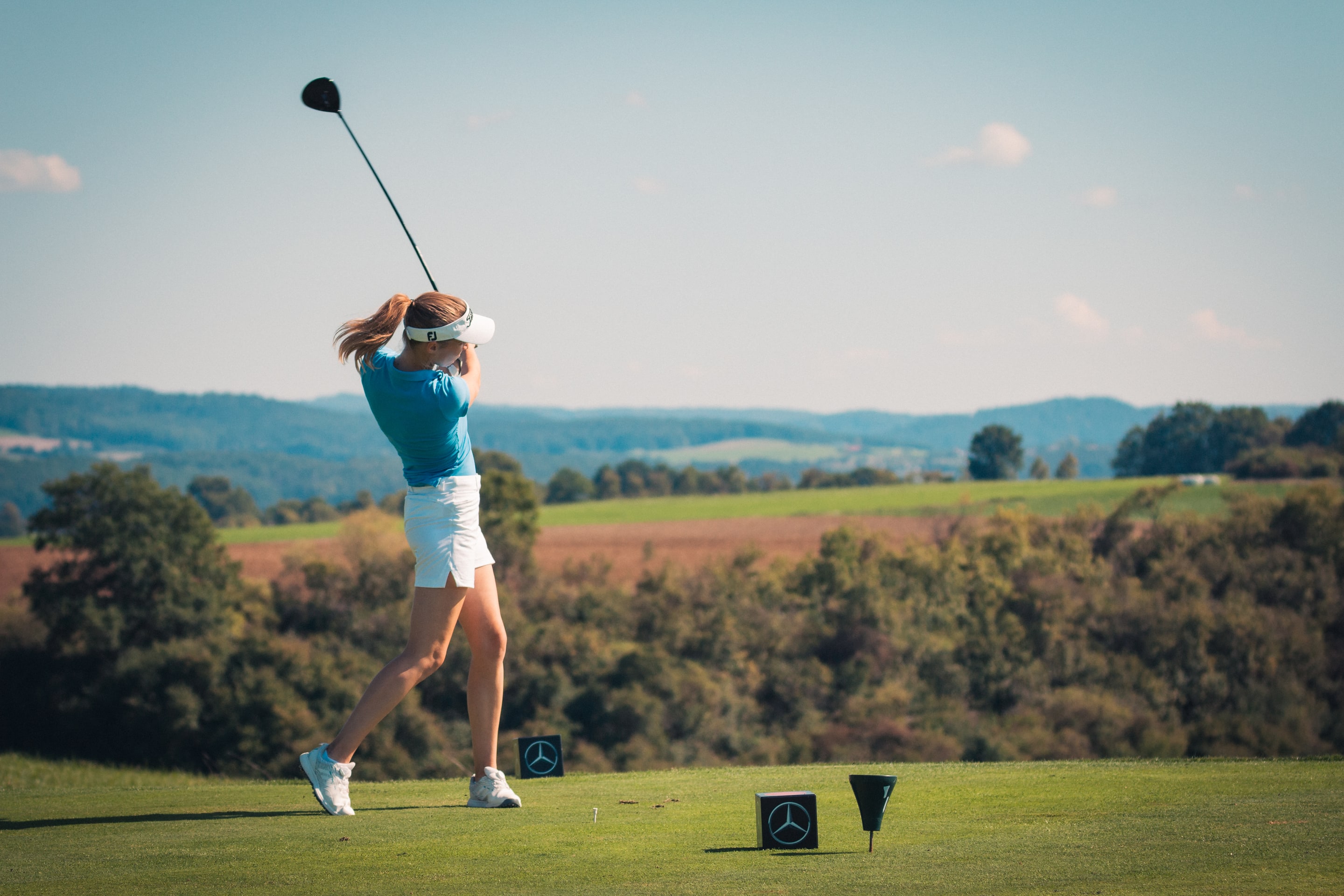 Golferin direkt nach dem Abschlag, den Schläger über die Schulter geschwungen, vor weiter Hügellandschaft mit Wäldern und Feldern